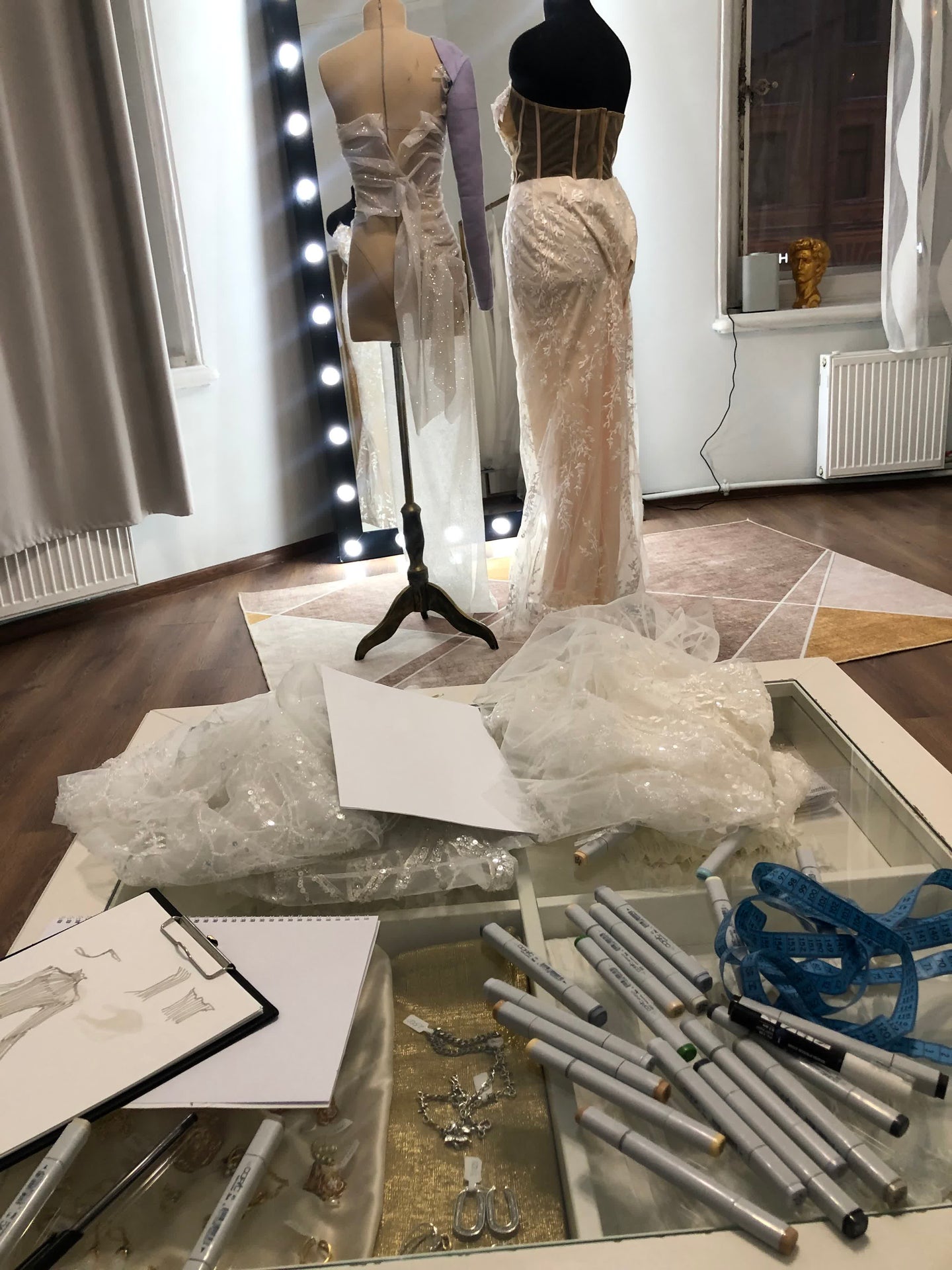 Atelier designer workspace with Copic markers, tape measure, dress sketches and dress forms with gowns in progress