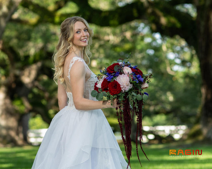 Hope on her wedding day, smiling and holding a cascading bouquet of burgundy roses and peonies at a beautiful outdoor Southern venue