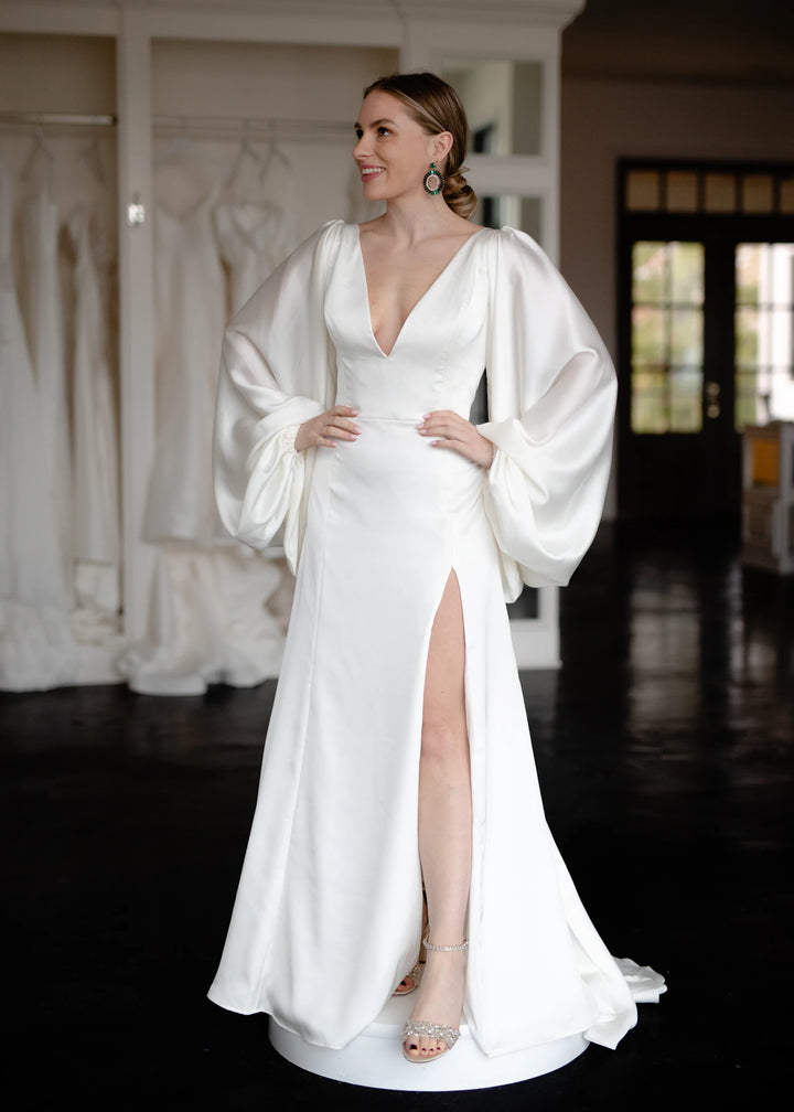A bride in a white satin wedding gown with puff sleeves standing in a bridal boutique, reviewing the fit in soft natural light