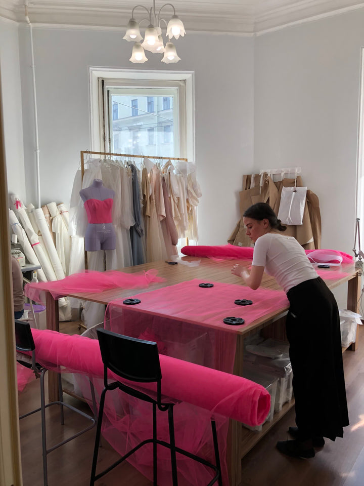 Seamstress cutting fabric at a large atelier worktable — a dress built once for one bride, the opposite of a rental garment reused across many